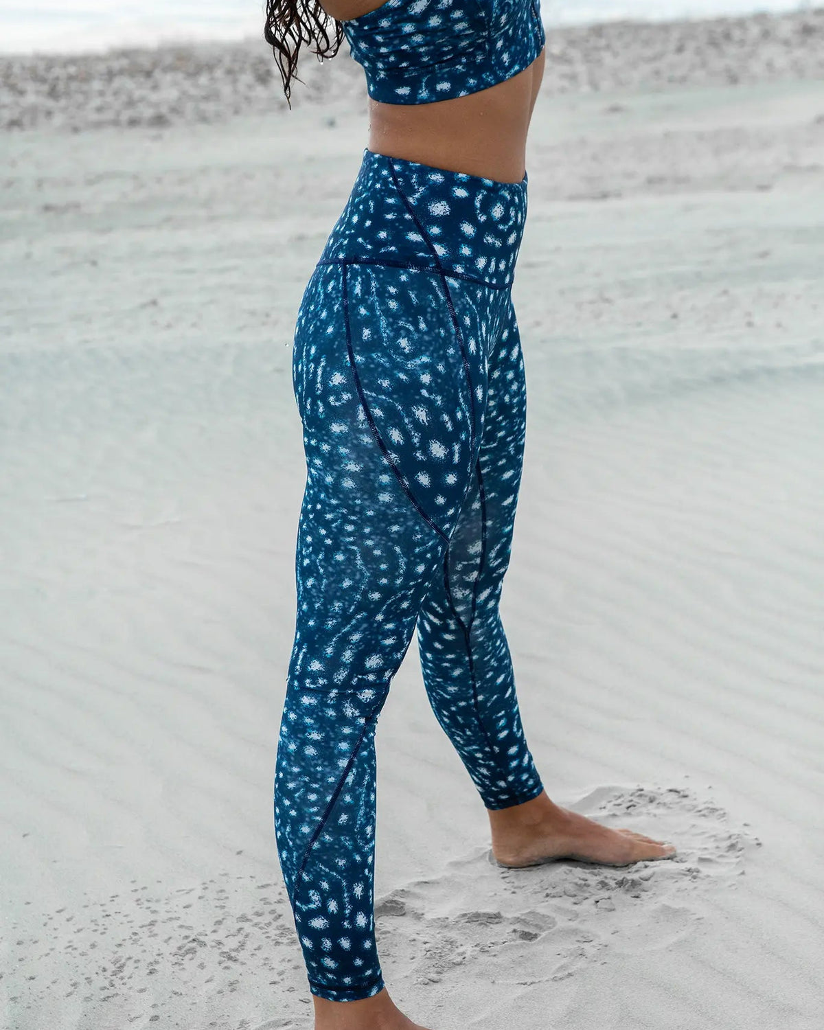 A person standing on a sandy beach wearing a matching NAUTRA's whale shark print sports bra and high-waisted leggings set.