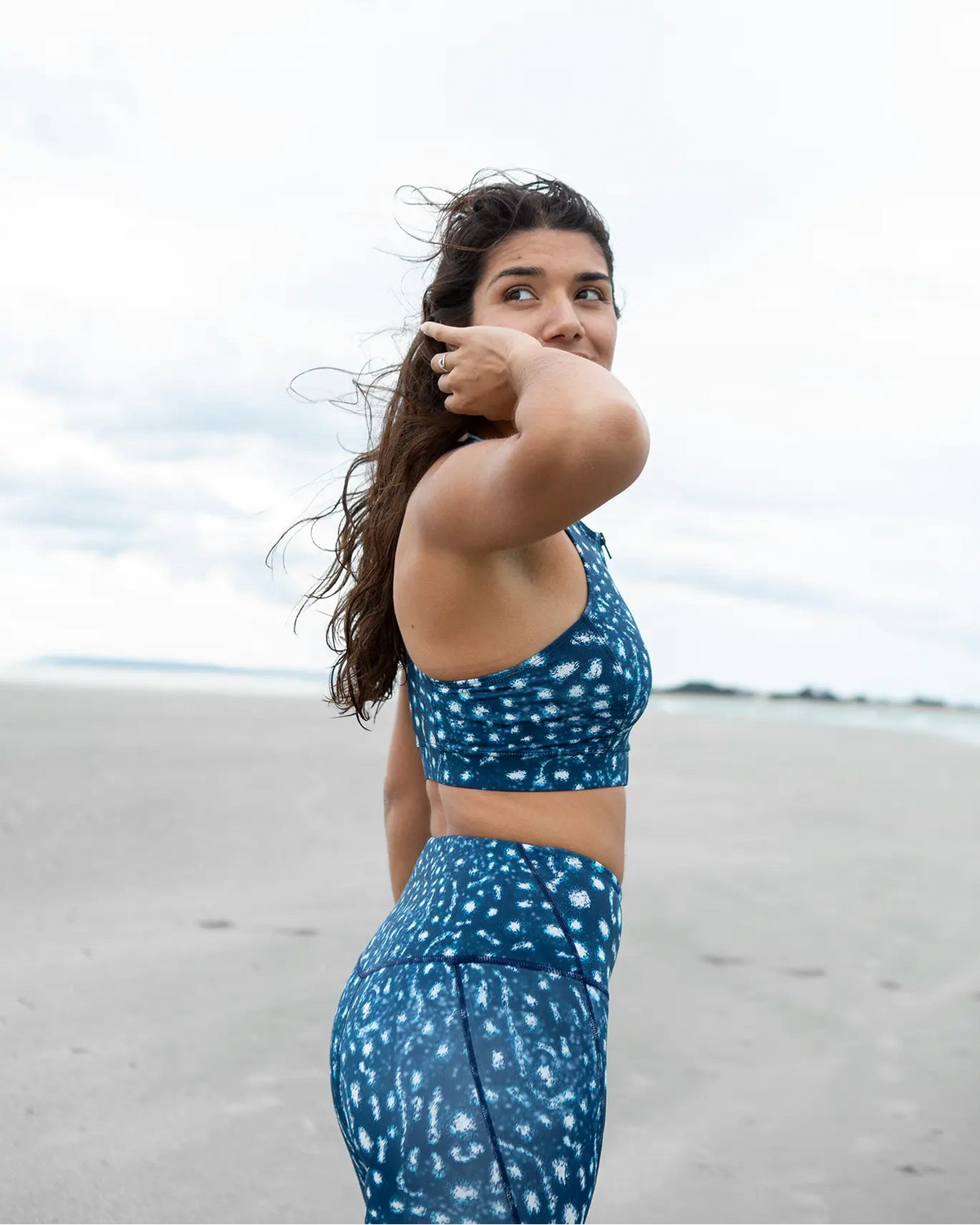 A woman standing on a beach wearing NAUTRA's whale shark print activewear set consisting of a sports bra and high-waisted leggings.