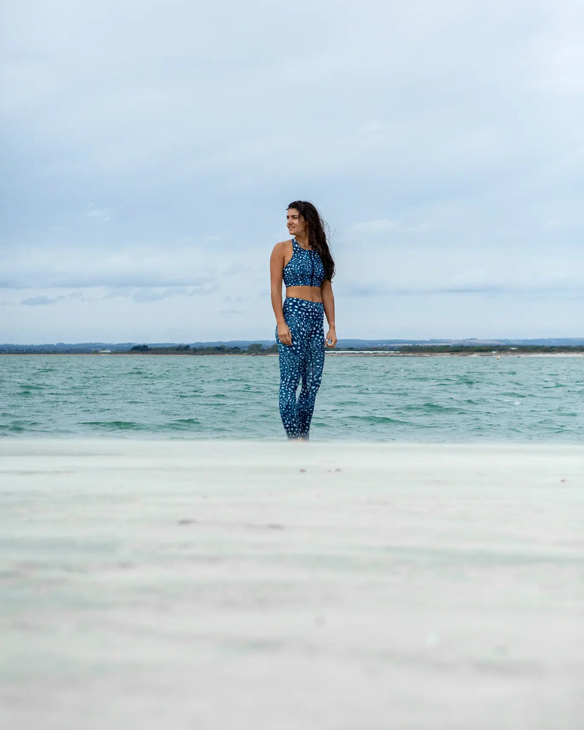 A woman wearing NAUTRA's whale shark print two-piece activewear set stands in shallow water on a beach under a cloudy sky.