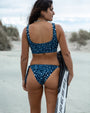 A woman on a sandy beach wearing NAUTRA's whale shark Scoop Sports Bra and Tie Side Bikini Bottoms, holding  Cressi fins.