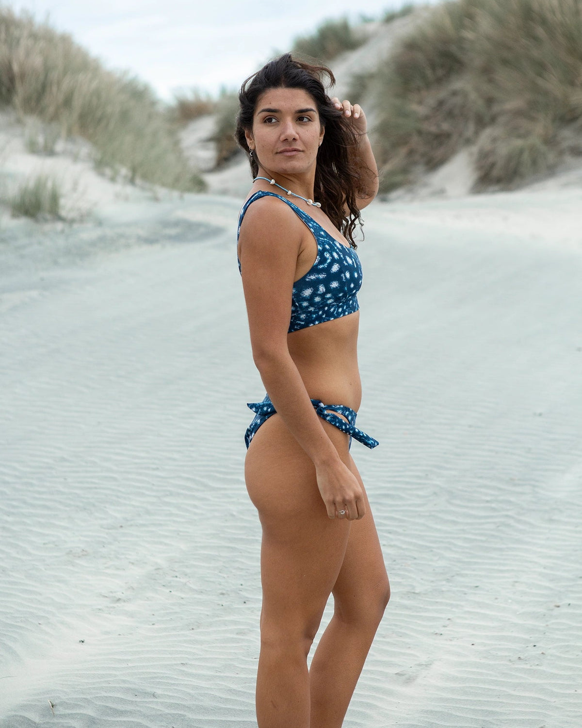A woman wearing NAUTRA's whale shark Scoop Sports Bra and Tie Side Bikini Bottoms stands on a sandy beach with dunes and grass in the background.