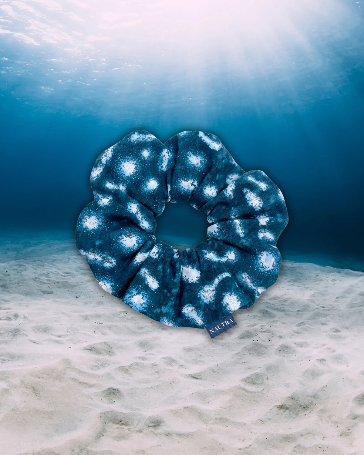 NAUTRA whale shark hair tie scrunchie, floating on a sandy ocean floor with sun rays filtering down from the water surface above.