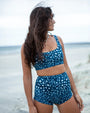 A woman standing on a beach wearing NAUTRA's whale shark print high waisted surf shorts and matching scoop bikini bra. The image is shot from behind, showing her profile and the ocean in the background.