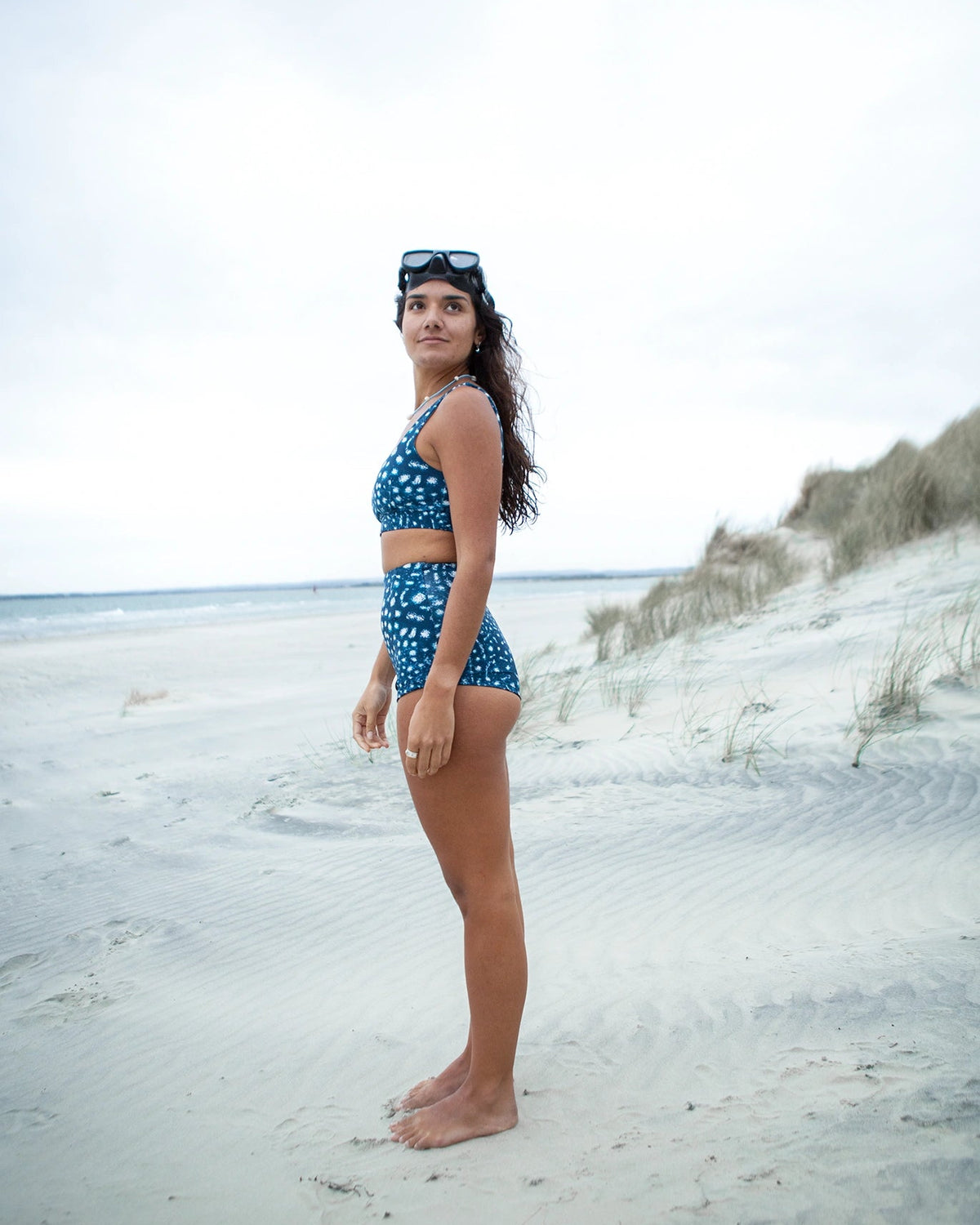 A woman stands on a sandy beach wearing NAUTRA's whale shark print high waisted surf shorts and matching scoop bikini bra with a black dive mask on her head, with dunes and ocean in the background.