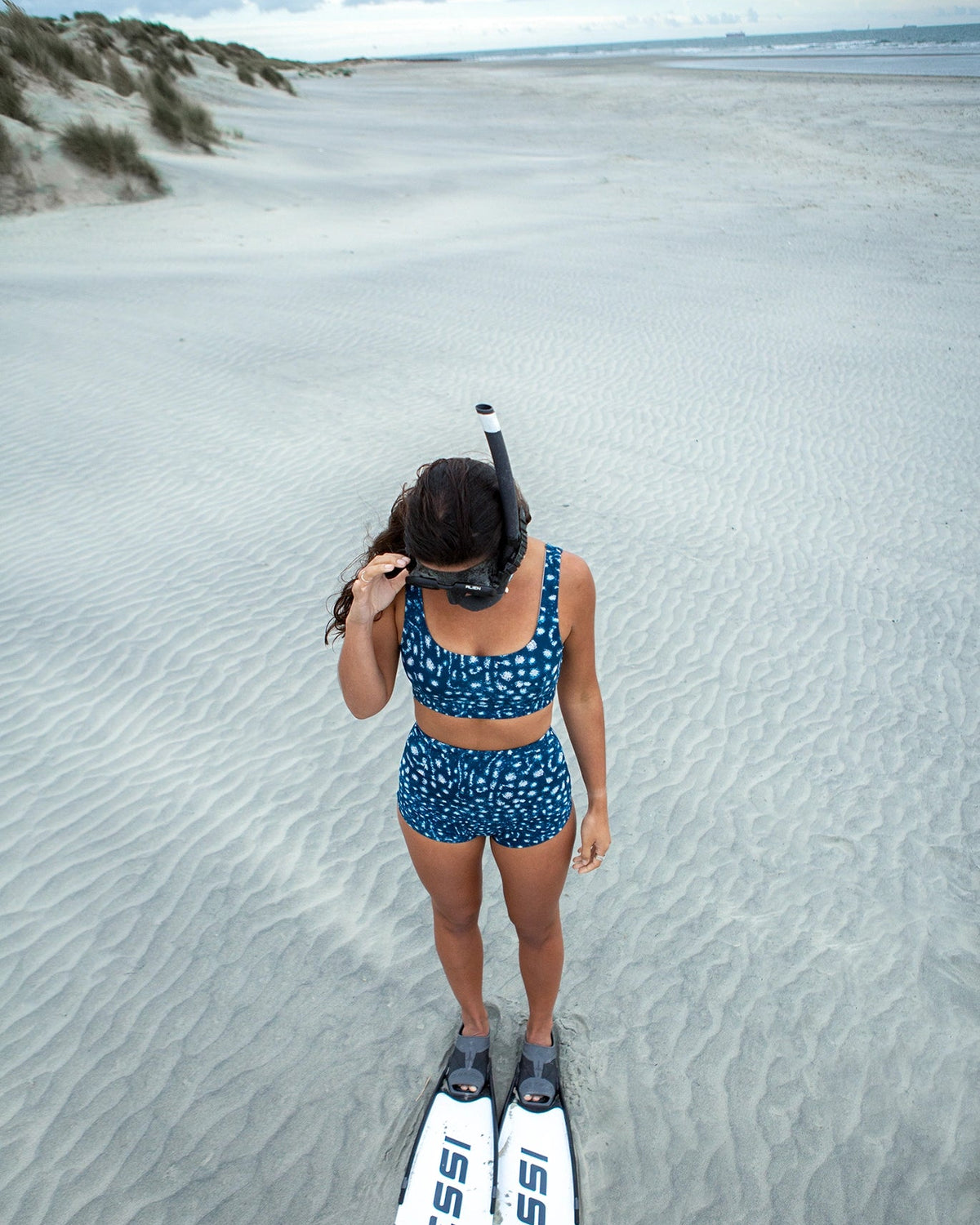 A woman stands on a sandy beach wearing NAUTRA's whale shark print high waisted surf shorts and matching scoop bikini bra, a snorkel mask, and white Cressi swim fins.