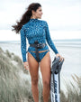 A woman stands on a beach dune holding a black and white board, wearing NAUTRA's whale shark print long sleeve rashguard and matching bikini bottoms with white spots and a black waist belt.
