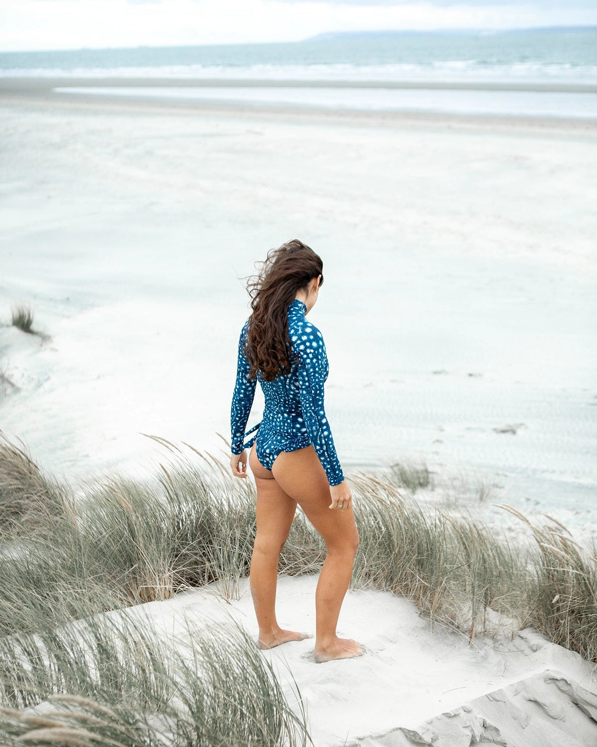A woman wearing NAUTRA's whale shark print long sleeve rashguard and matching bikini bottoms walks down sandy beach dunes toward the ocean.