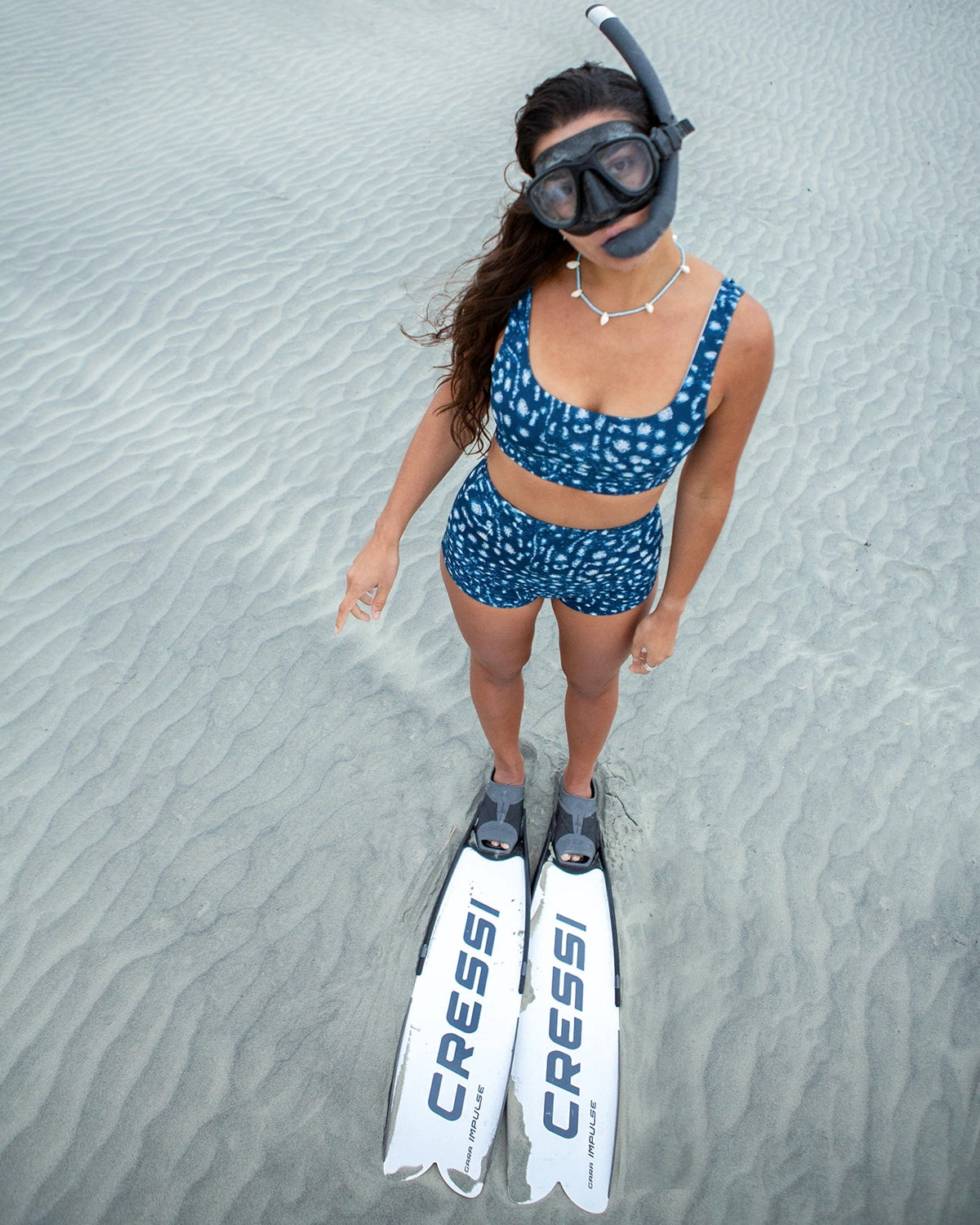 A woman stands on sand wearing NAUTRA whale shark scoop sports bra and high waisted surf shorts, a snorkel mask, and white Cressi flippers. The Cressi brand name is clearly visible on the flippers.