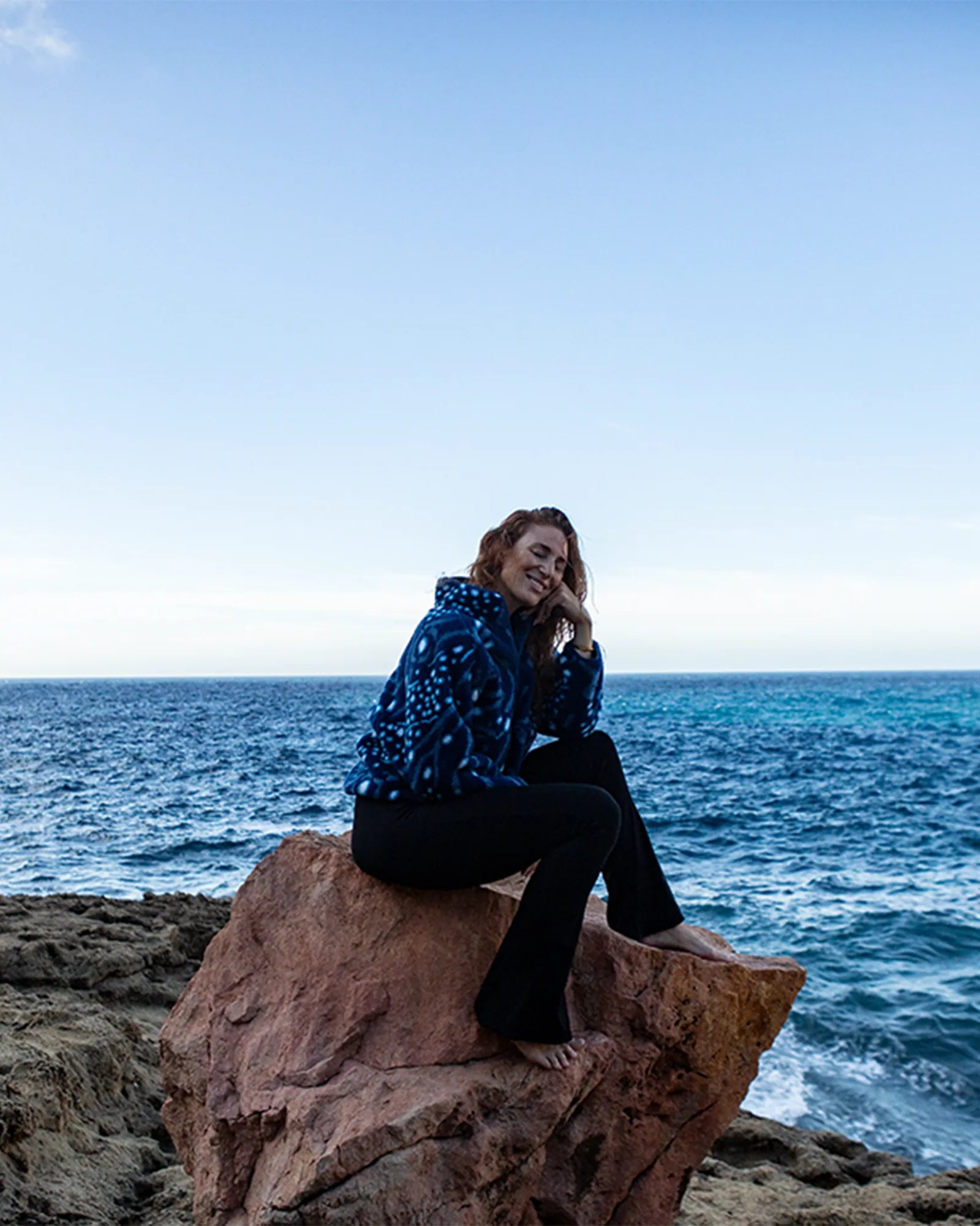 Woman sitting on coastal rock wearing Whale Shark recycled sherpa fleece by the sea.